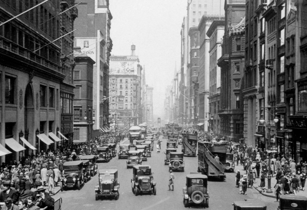 Image historique en noir et blanc d'une rue animée, peuplée de piétons et de voitures.